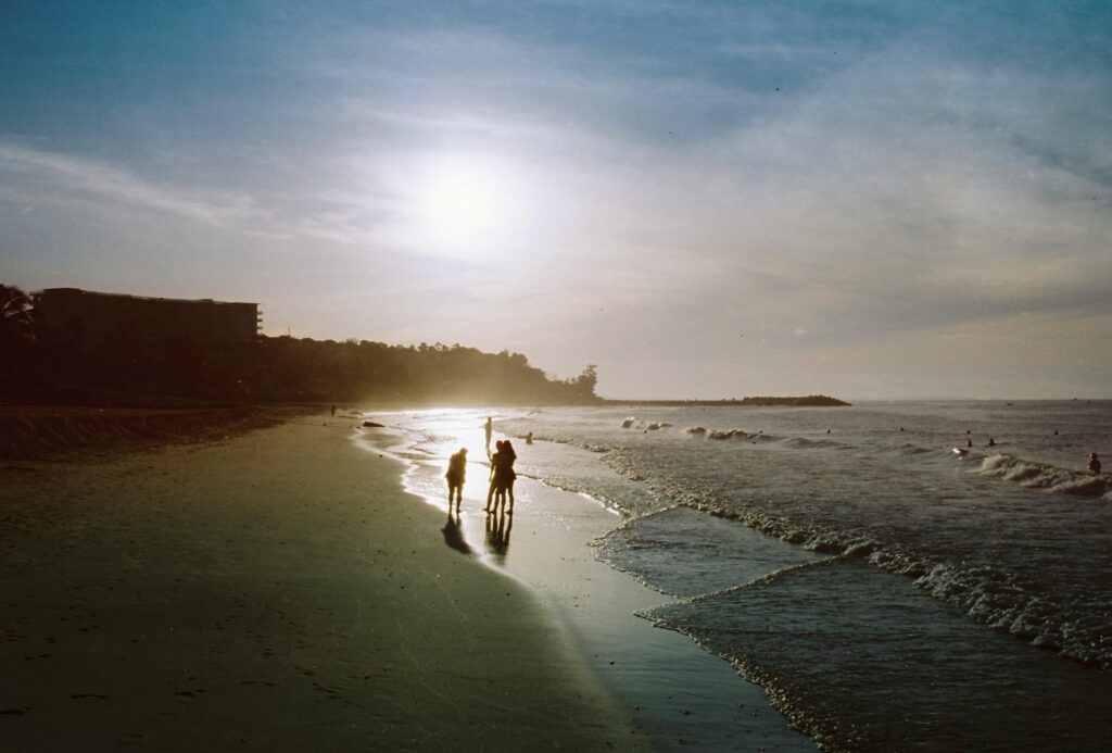 A serene beach scene with people walking by the ocean at sunrise, casting long shadows.