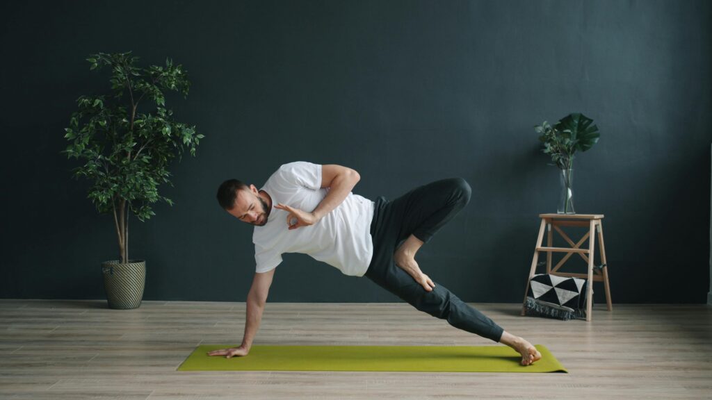 Adult male performing a side plank yoga pose indoors on a green mat.