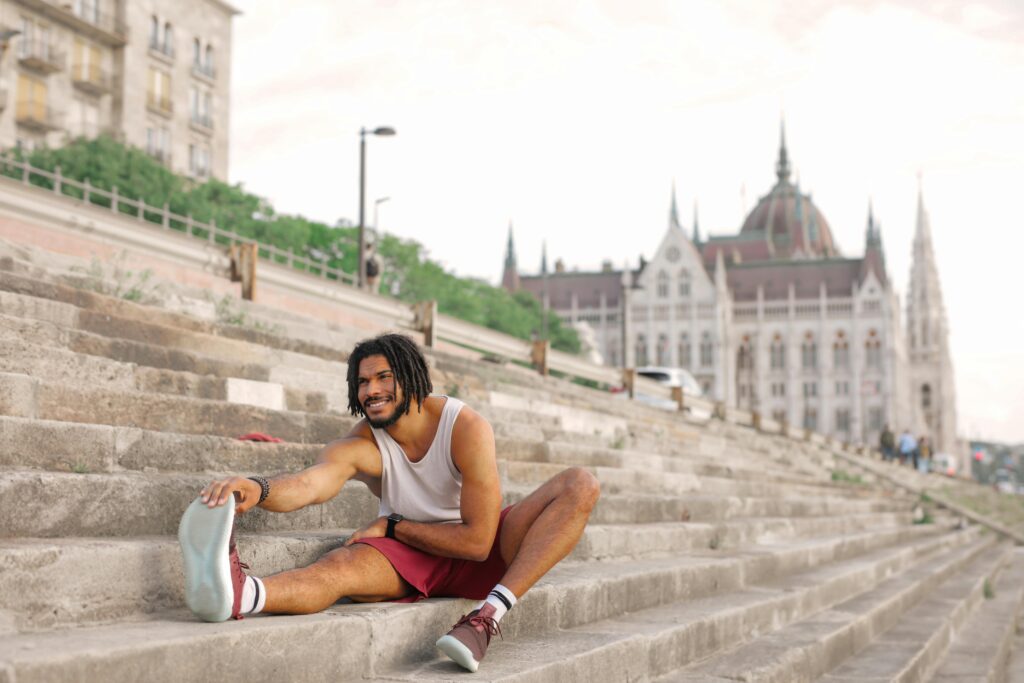 Man with dreadlocks smiling while stretching on city steps near architectural landmark.