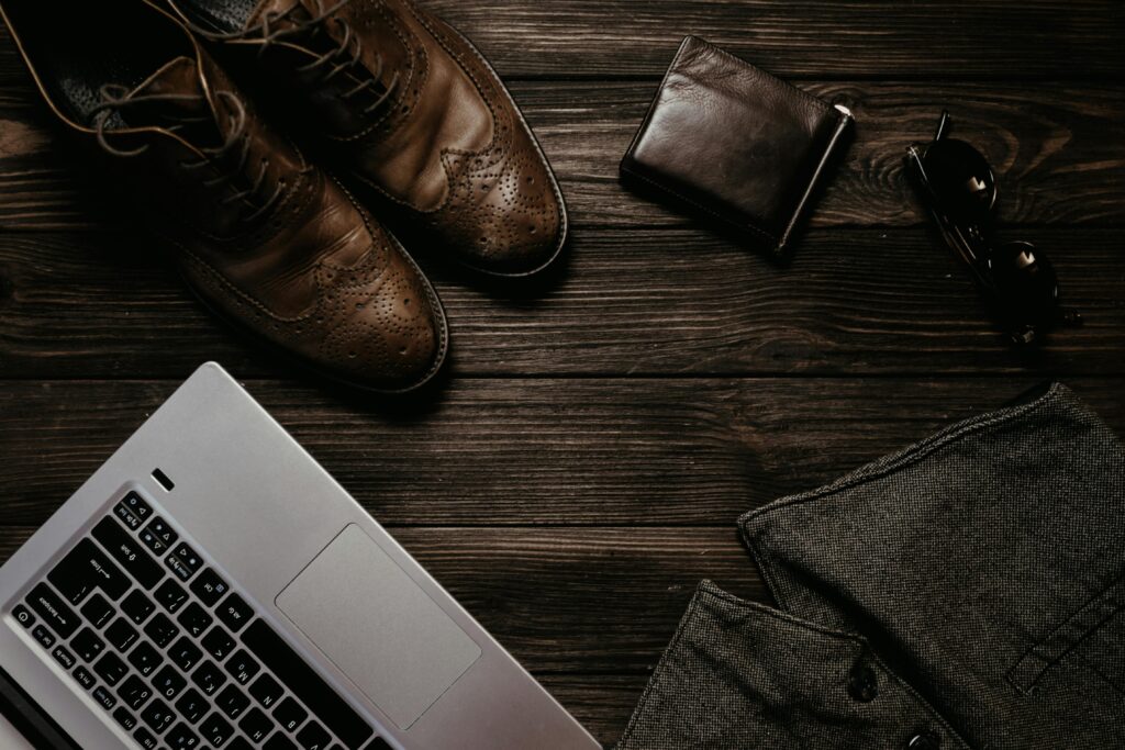 Modern workspace featuring laptop, leather shoes, wallet, and sunglasses on a wooden desk.