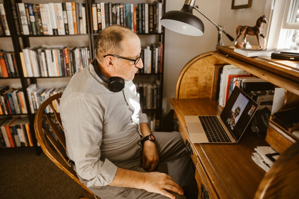 Senior adult man video calling from home office desk, surrounded by books, emphasizing connectivity and technology.