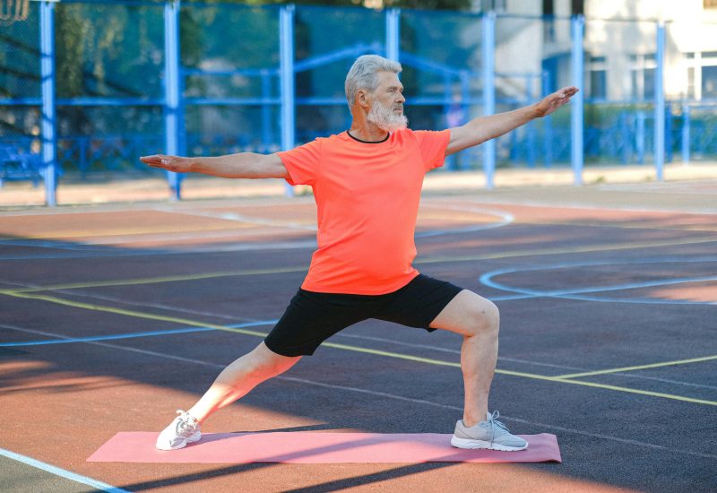 Elderly man performing warrior II yoga pose on an outdoor court.